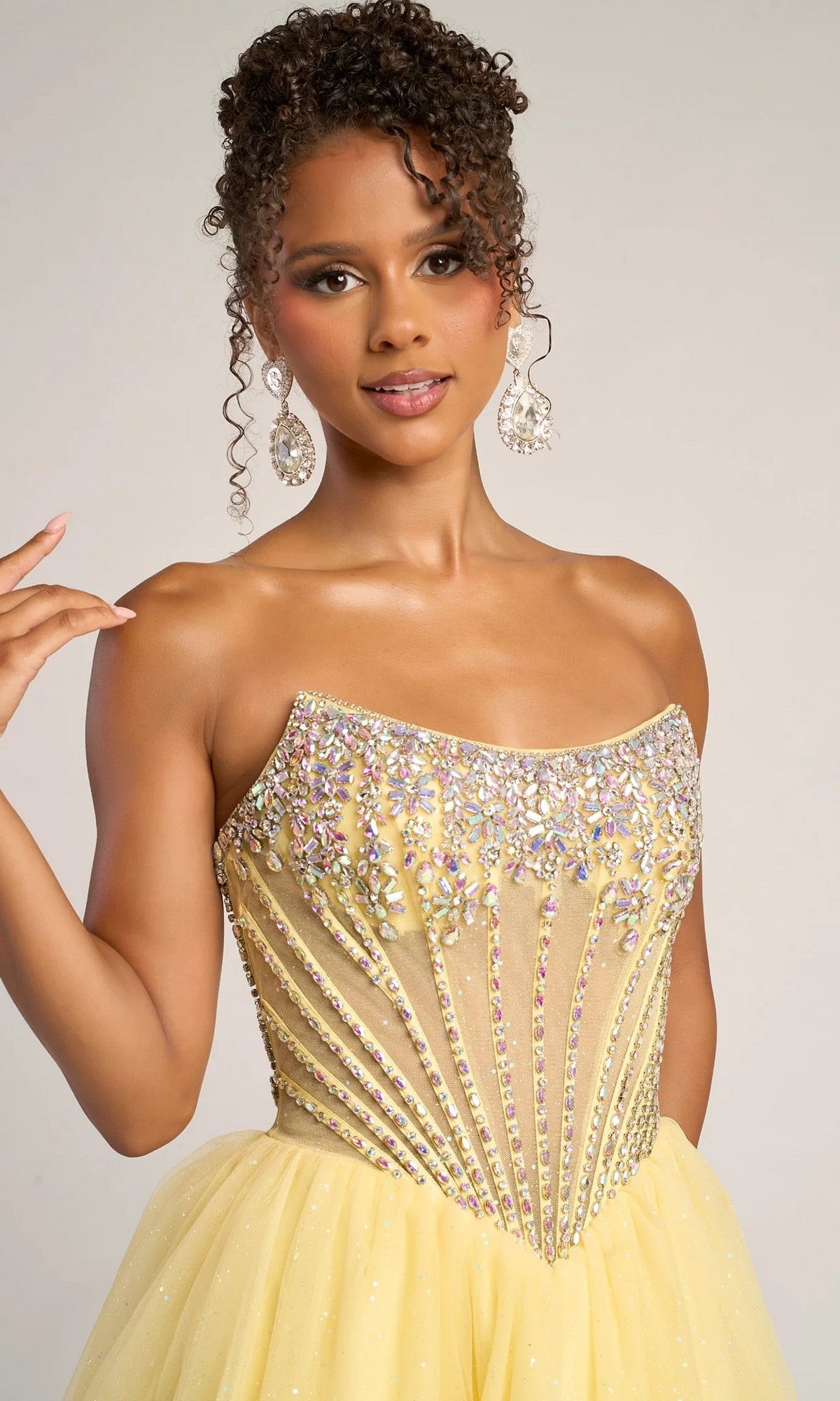 A woman poses against a plain background in the Portia and Scarlett XO047 long formal dress, featuring a strapless tulle ball gown design with intricate rhinestone embellishments on the beaded corset bodice and large dangling earrings.